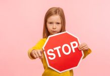 Portrait of cute serious bossy little girl holding Stop symbol, showing red traffic sign and pointing to camera, warning you about road safety rules. indoor studio shot isolated on pink background