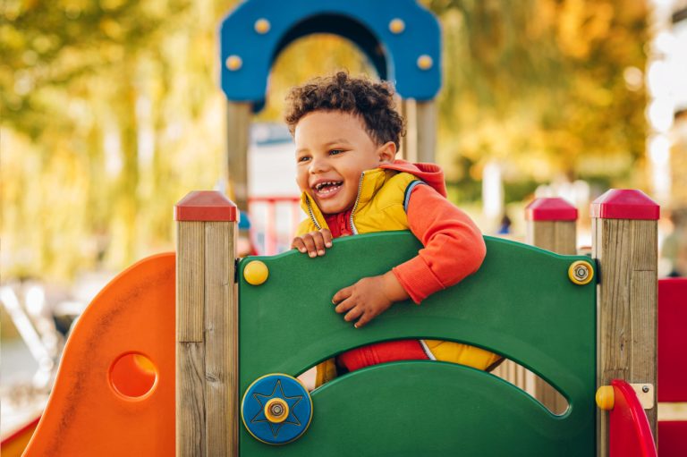 Adorable little 1-2 year old toddler boy having fun on playground, child wearing orange hoody jacket and yellow vest
