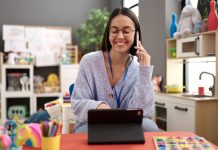 Young beautiful hispanic woman working as a teacher talking on smartphone using touchpad at kindergarten