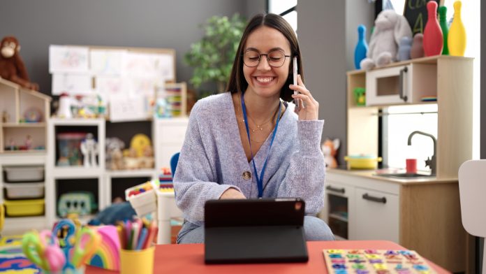 Young beautiful hispanic woman working as a teacher talking on smartphone using touchpad at kindergarten