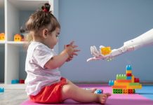 Baby girl playing with multi colored toy blocks while she is trying to grab a piece of blocks from humanoid robotic hand. She is sitting on the floor. Shot indoor with a full frame mirrorless digital camera.