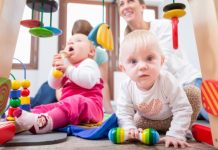 Cute baby girl showing progress and curiosity by trying to reach multicolored wooden toys, while sitting on the floor with her mother and a family friend