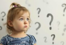 Cute little toddler girl with curious and thoughtful face expression, standing in the room in front of the wall full of question mark symbol drawings, wondering concept, child learning and education