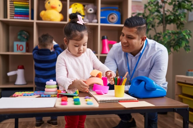 Vroege kinderopvang heeft langdurig positief effect op schoolprestaties in Noorwegen Mannelijke pedagogisch professional zit aan een tafel met jong meisje die speelt met speelgoed.