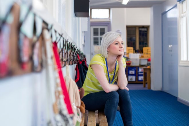 Wat kunnen we leren van uitstromers in de kinderopvangbranche? Young, female teacher sitting on a bench in the cloakroom of school. She is looking pensively out of the window.