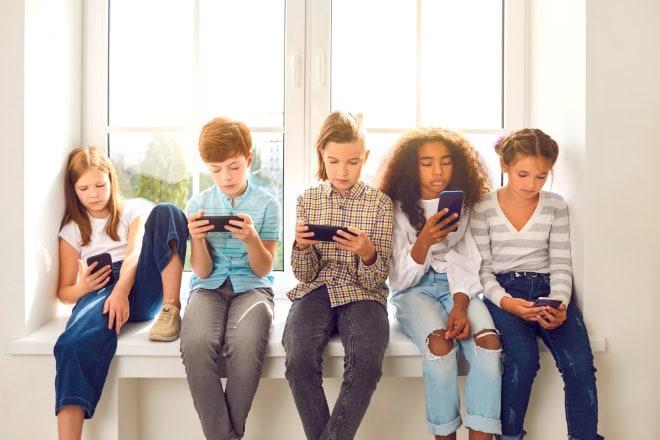 AdobeStock_412273512-2 Children sitting on windowsill, playing online games on mobile phones and ignoring each other