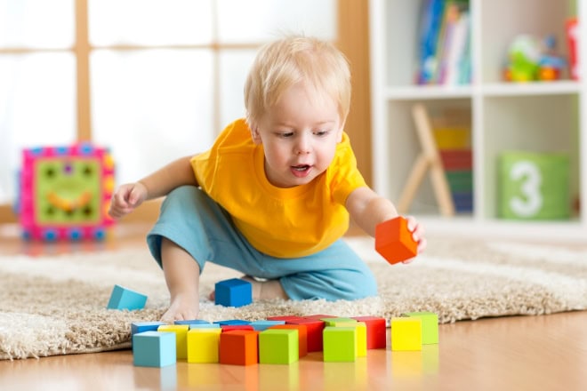 AdobeStock_76035644 kl Jong kindje met blond haar in geel t-ahirt speelt met blokken in knallende kleuren.