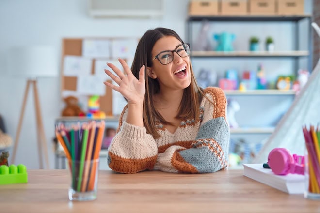 Young beautiful teacher woman wearing sweater and glasses sitting on desk at kindergarten Waiving saying hello happy and smiling, friendly welcome gesture welke-redenen-hebben-zzp'ers-om-te-vertrekken-ui-t-de-kinderopvang-of-te-blijven