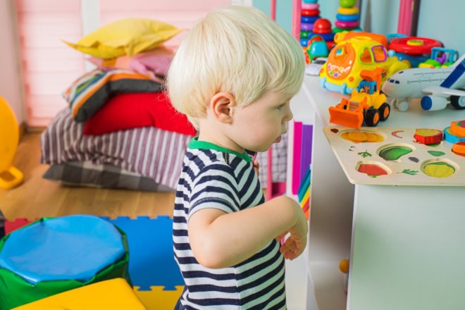 AdobeStock_407617322-2 Lieve peuterjongen in een gestreept T-shirt, staand bij een plank met speelgoed op de kleuterschool en wegkijkend. Het jongetje voelt zich onzeker tijdens zijn eerste dag op de kleuterschool.